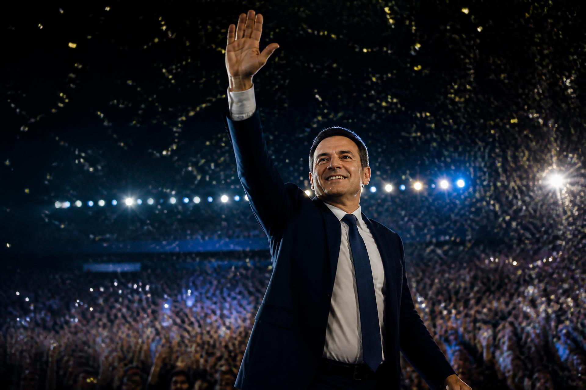 Liderança política fictícia em um palco de campanha acenando para o público em clima de celebração